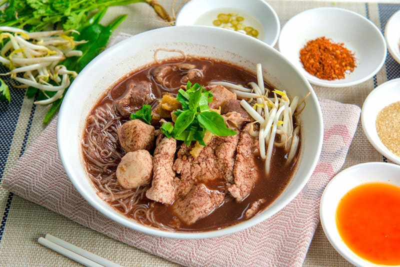 Bowl of dark Thai boat-noodle soup with pork slices, a pork ball, basil and bean sprouts, with condiments