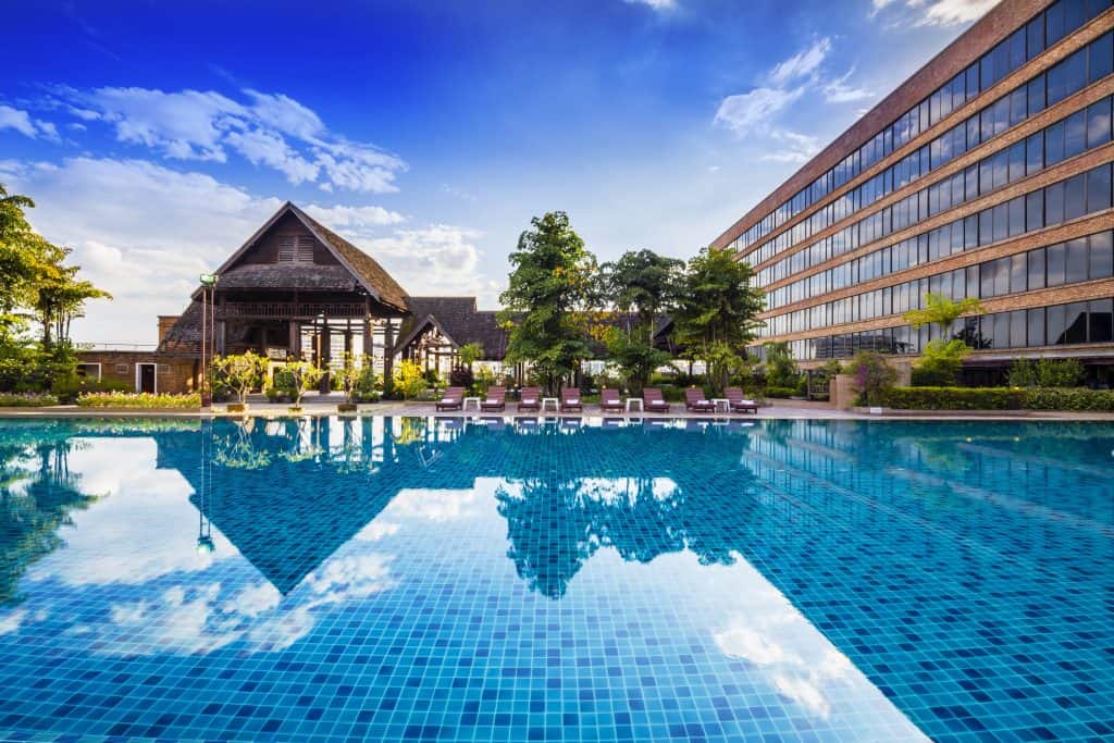 Large pool reflecting clouds between a wooden Lanna building and the Lotus Pang Suan Kaew hotel tower