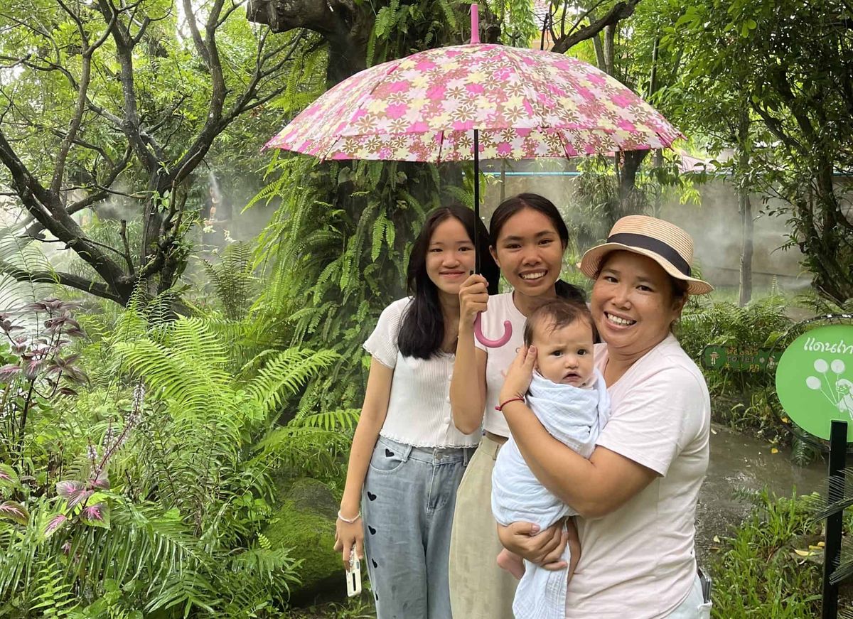 A smiling nanny holding a baby beside her two teenage daughters under a floral umbrella in a misted garden