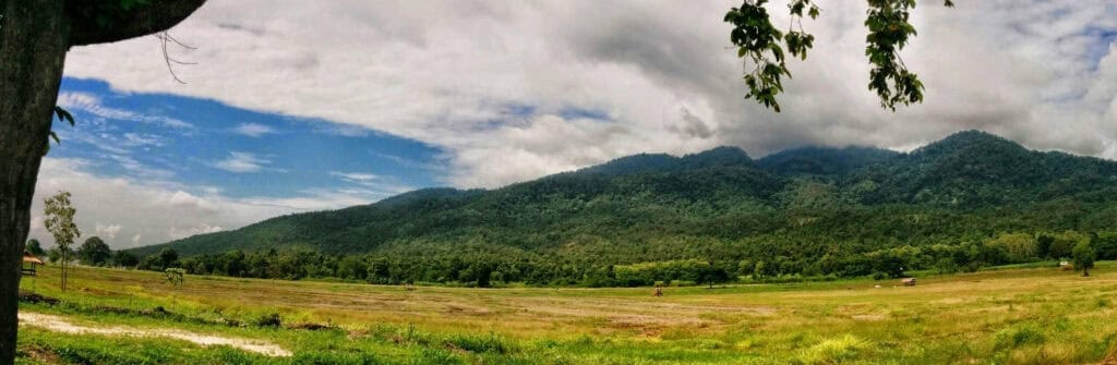 Panorama of a green Chiang Mai countryside field below forested Doi Suthep mountains under a cloudy sky