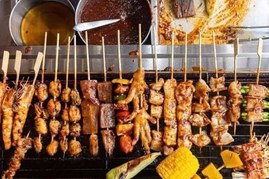 Rows of grilled meat and vegetable skewers over hot coals with mala dipping sauces and grilled corn