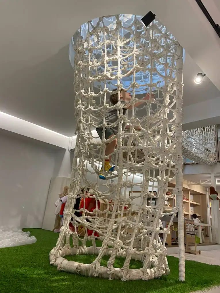 Child climbing a tall rope-net tower over turf and a ball pit at Wooden Ville Kids Cafe