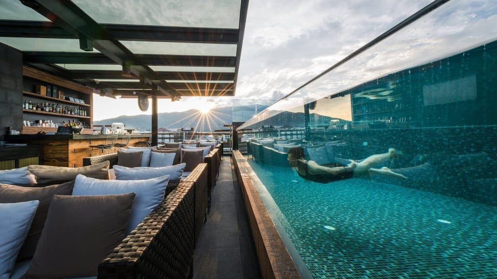 Akyra Rise Bar rooftop infinity pool at sunset, a swimmer behind the glass wall and rattan lounge seating