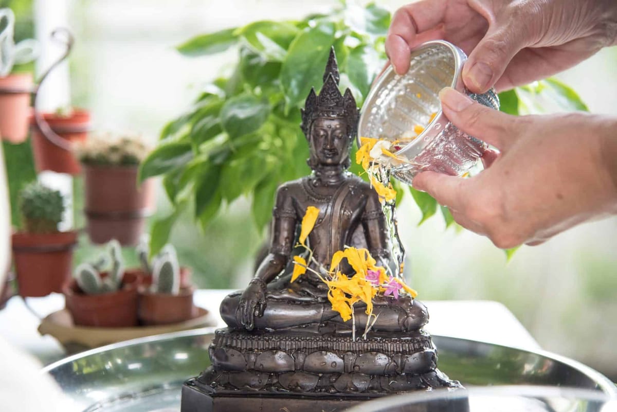 Hands pouring scented water and marigolds from a silver bowl over a bronze Buddha statue for Songkran, the Thai New Year