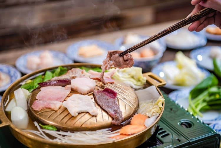 Chopsticks grilling pork on a brass moo kratha dome with a broth moat and side dishes of vegetables