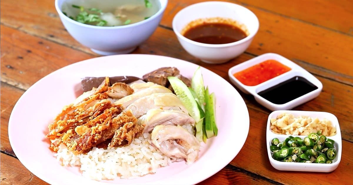 Plate of khao man gai chicken rice with steamed and fried chicken, cucumber, broth and dipping sauces