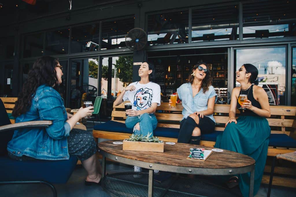 Four women laughing over drinks on bench seating at an open-air bar