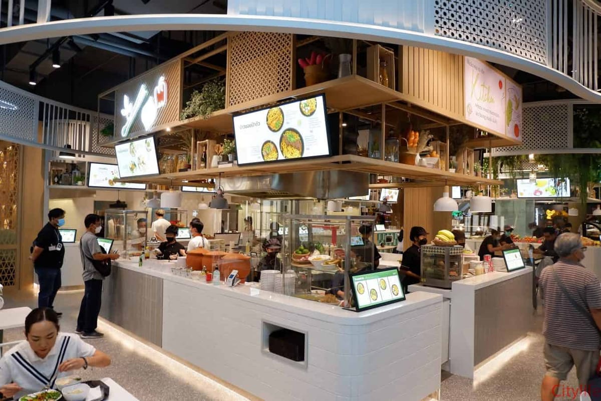 Modern shopping-mall food court with a central counter, menu screens and masked staff serving customers