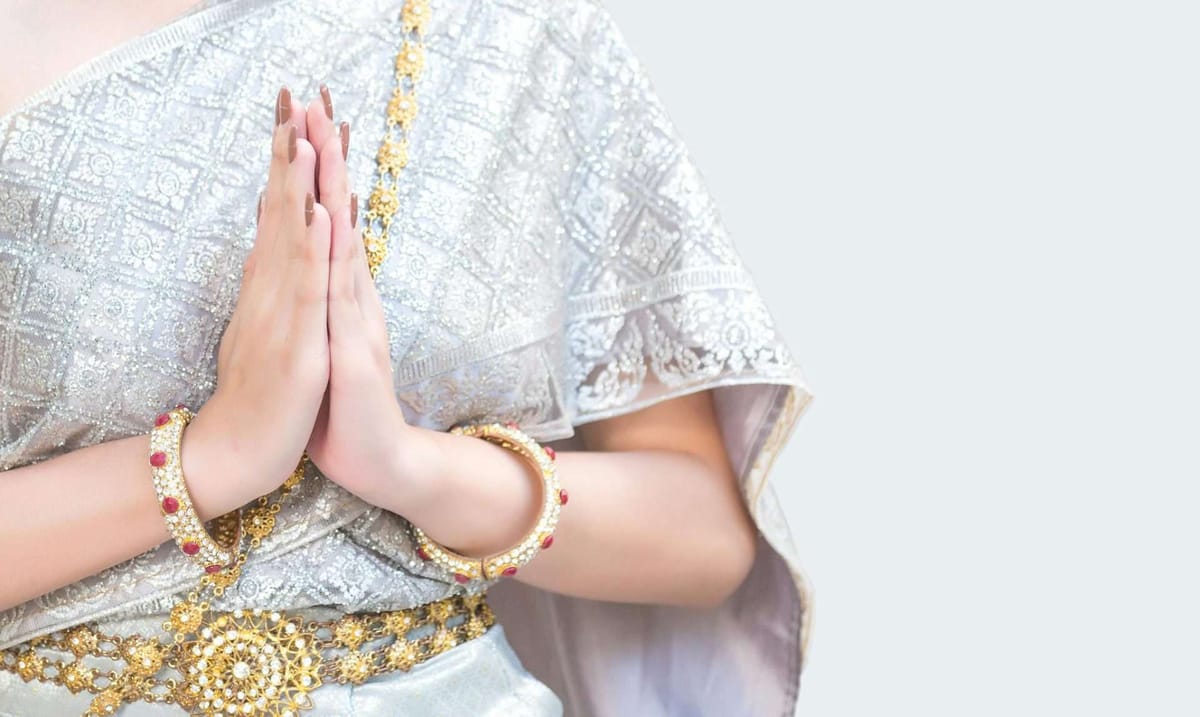 Woman in a traditional silver Thai dress and gold jewelry making a wai greeting