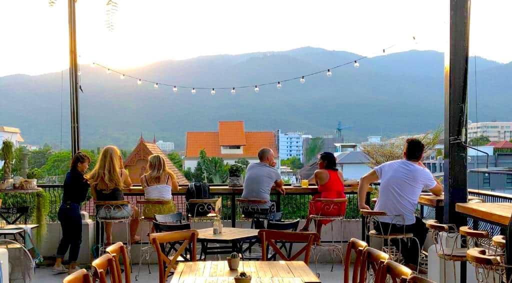Patrons at a Nimman rooftop bar watching the sunset over the mountains beneath string lights