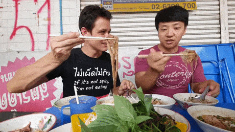 Two men slurping noodles at a Thai street-food stall piled with bowls and herbs