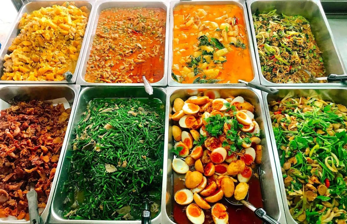 Metal buffet trays of Thai curries and stir-fries behind glass at a khao rad gang curry-over-rice shop
