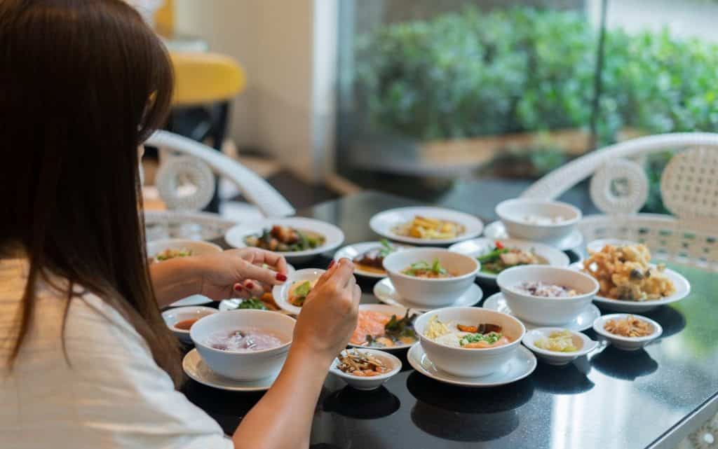 Woman eating family-style from a spread of small Thai side dishes and rice bowls at a khao tom baat eatery