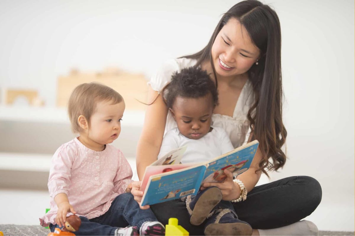 A young caregiver reading a picture book to two toddlers seated on the floor