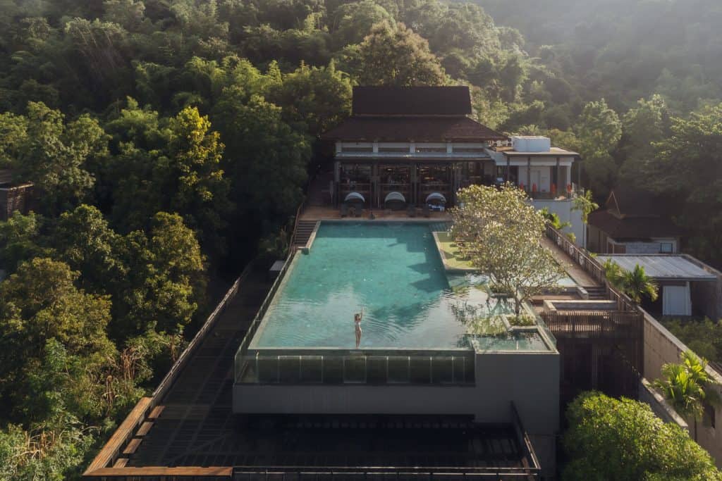 Aerial view of Veranda High Resort's hillside infinity pool ringed by forest, a woman at the edge