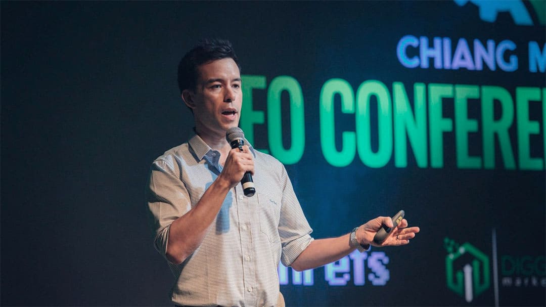 Speaker in a checked shirt addressing the audience with a microphone on stage at the Chiang Mai SEO Conference