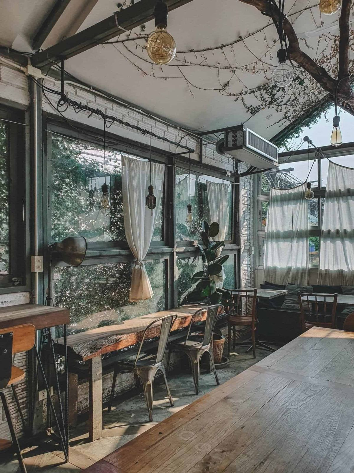 Rustic-industrial interior at The Barn Eatery & Design with reclaimed-wood tables and metal chairs under Edison-bulb pendants by garden windows