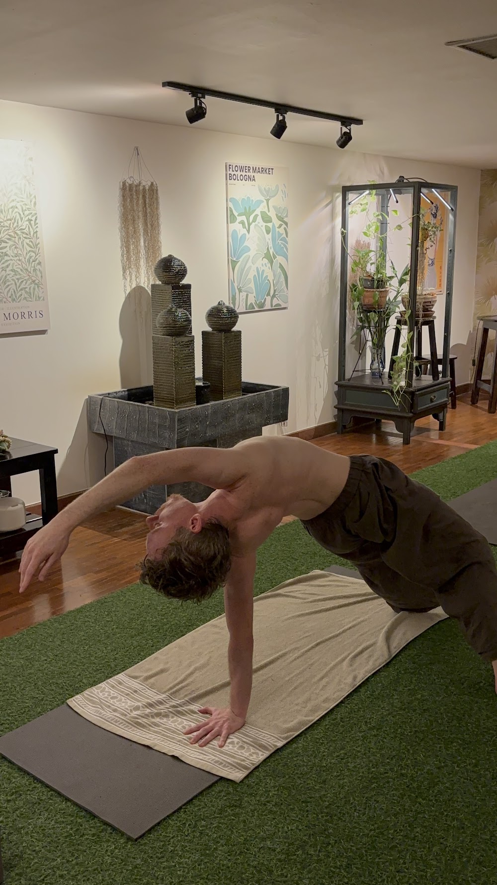 A man in a backbend yoga pose on a mat over artificial grass in Bliss's plant-filled studio room
