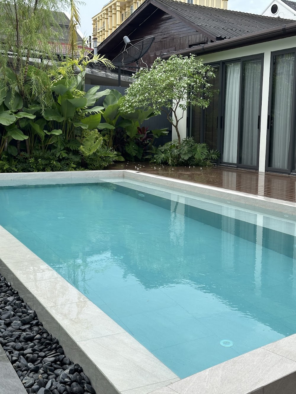 A turquoise rectangular pool with stone coping and pebble border beside the glass-doored building at OUR space