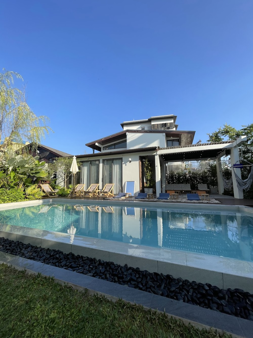 OUR space's white two-story villa behind the pool with deck chairs, blue loungers, a pergola and hammocks