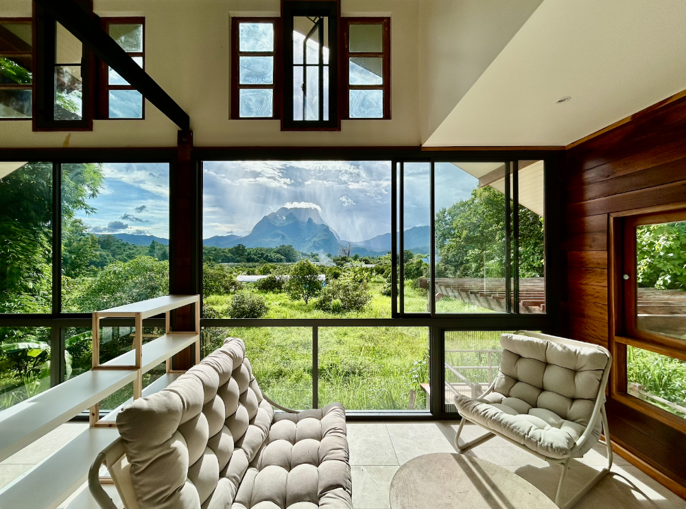 Indoor lounge with floor-to-ceiling windows framing the Chiang Dao mountain, cushioned tufted chairs and greenery outside