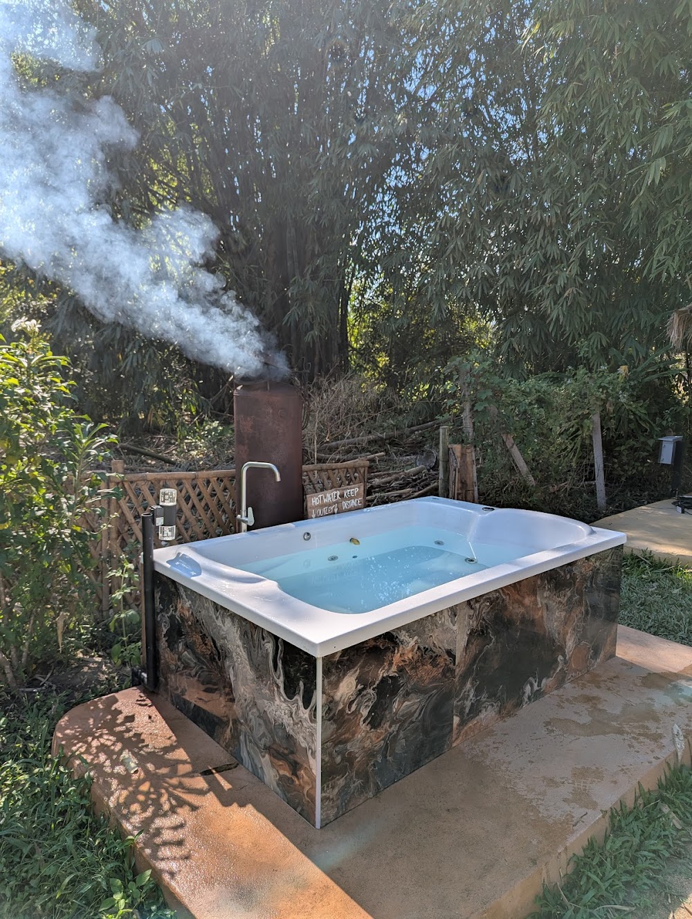 Outdoor marble-patterned hot tub steaming beside a wood-fired water heater in a leafy garden