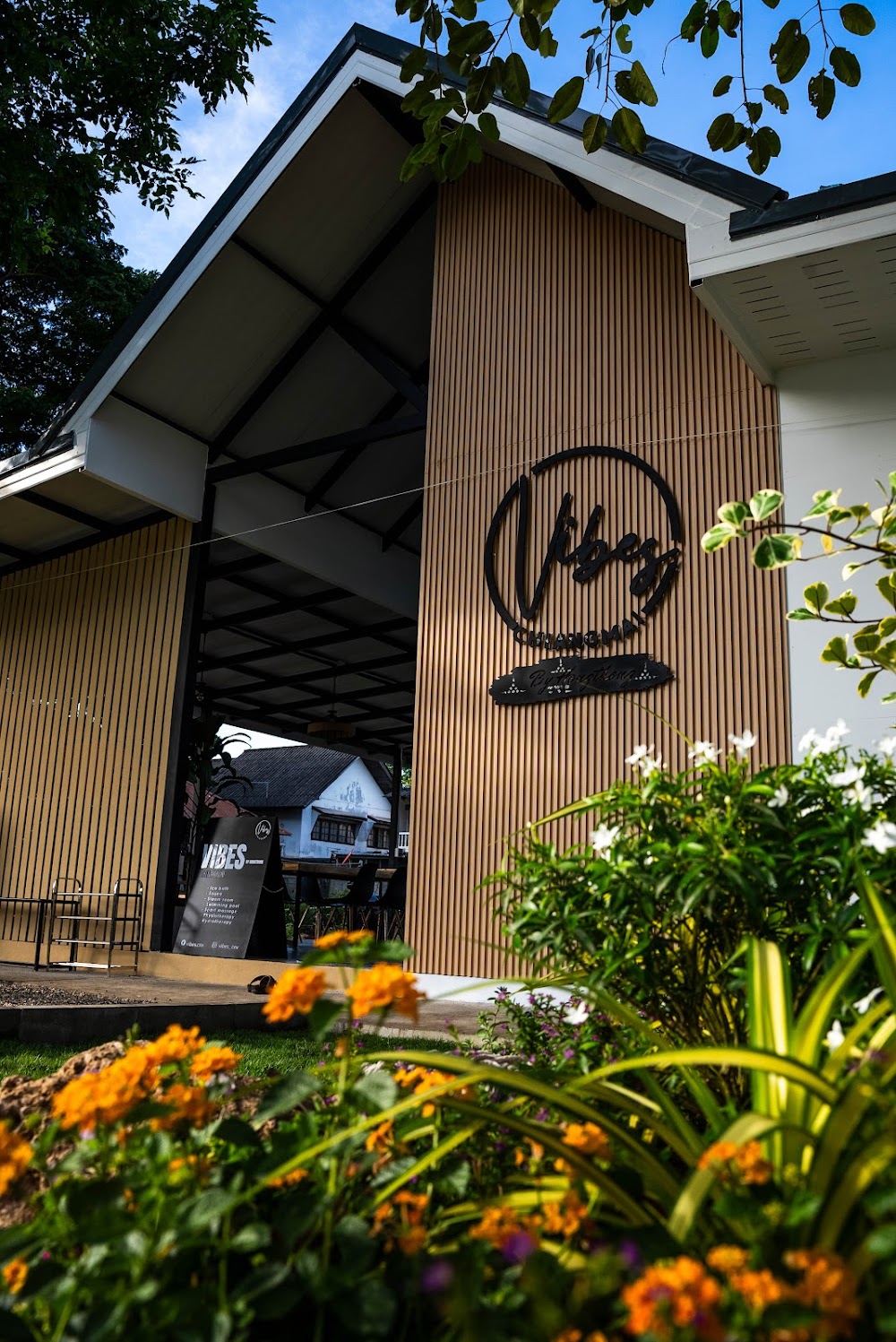The wood-slatted gable entrance of Vibes Chiangmai with its round logo above a flower garden