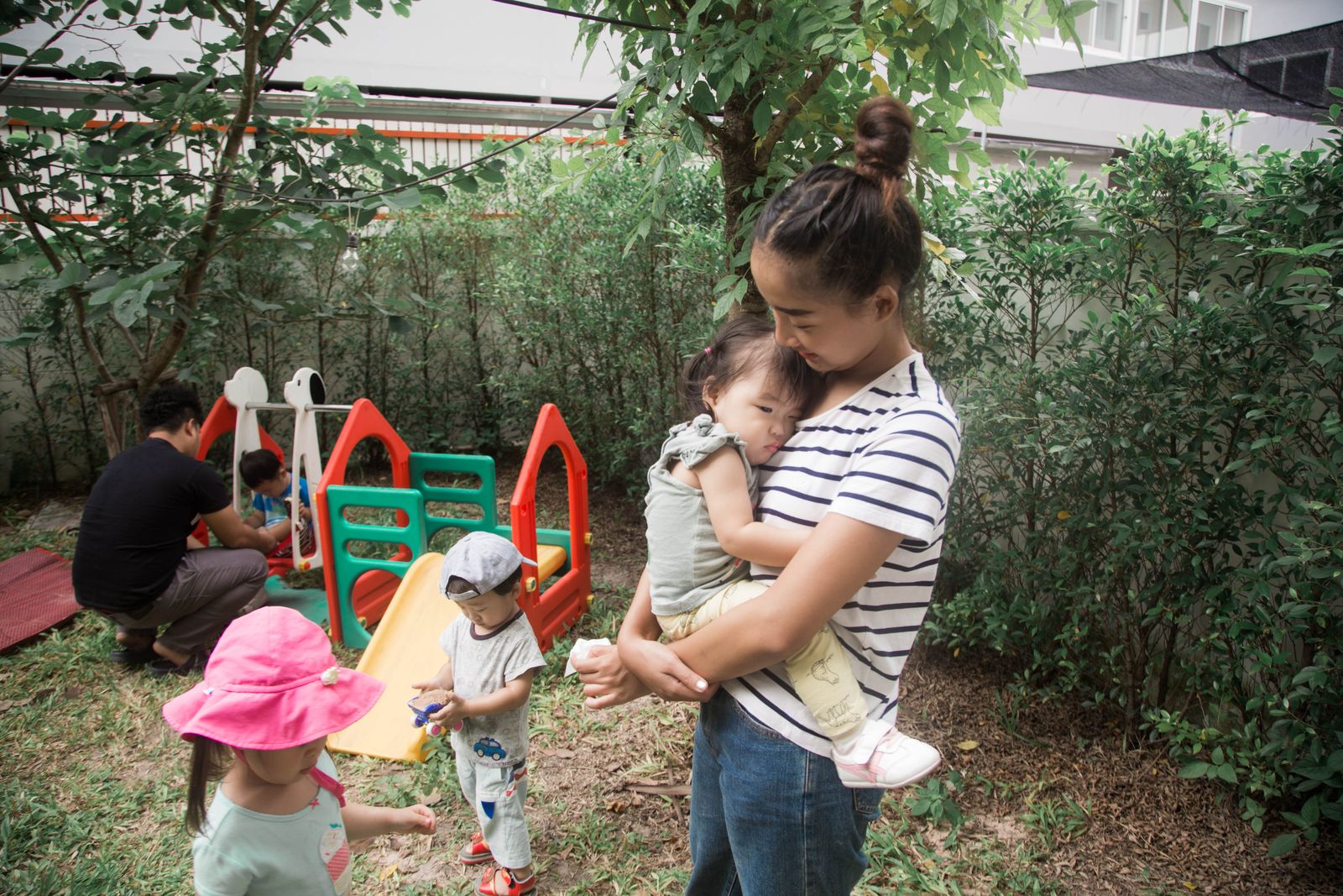 A caregiver holding a baby while young children play in a tropical garden